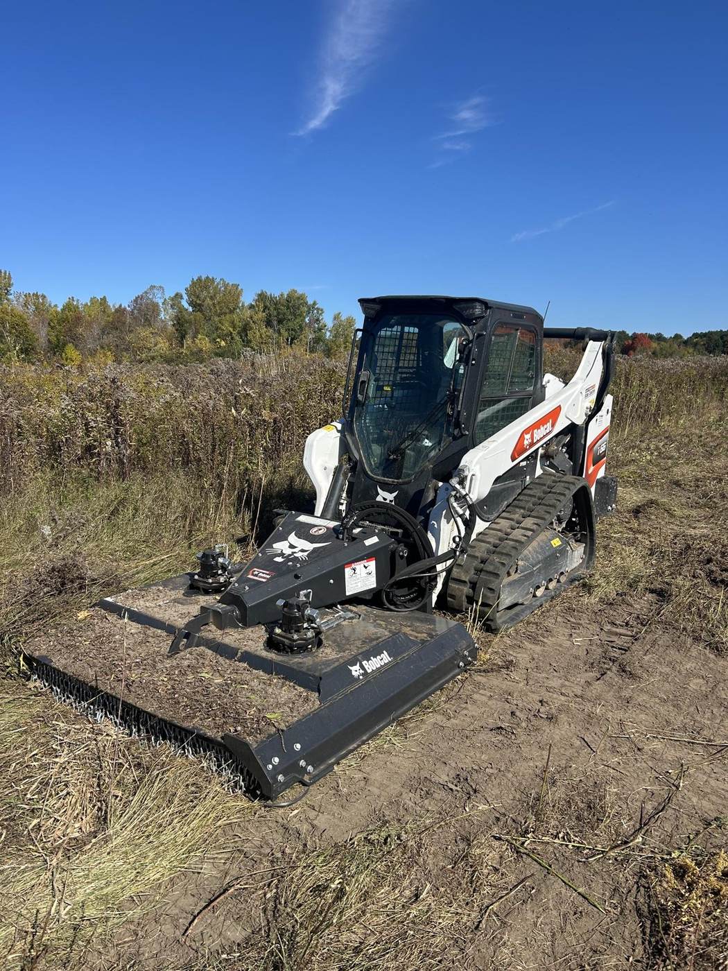 White Bobcat track loader using a brush cutter to clear an overgrown field